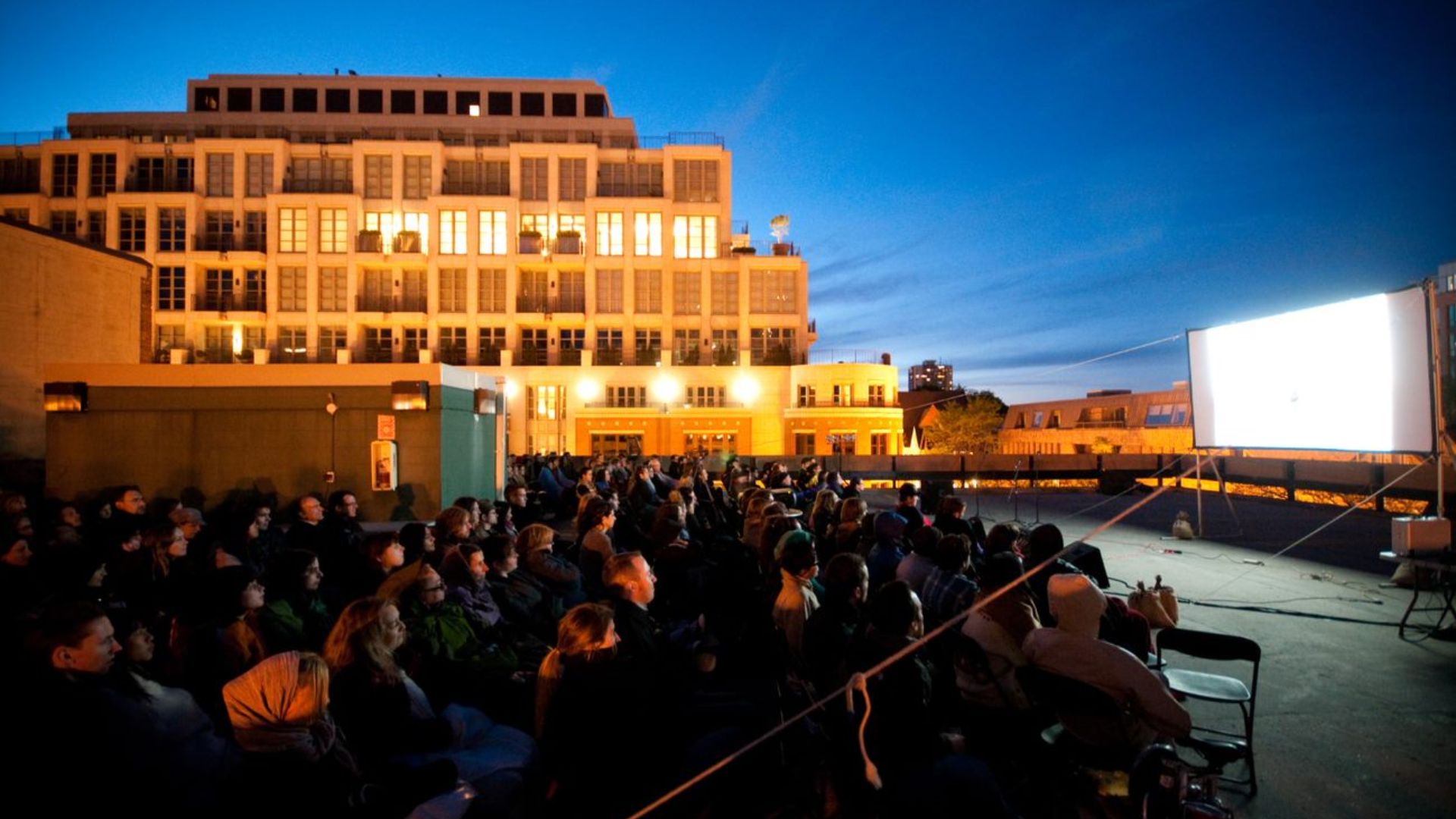 Outdoor film screening with a full audience on a building rooftop at dusk.