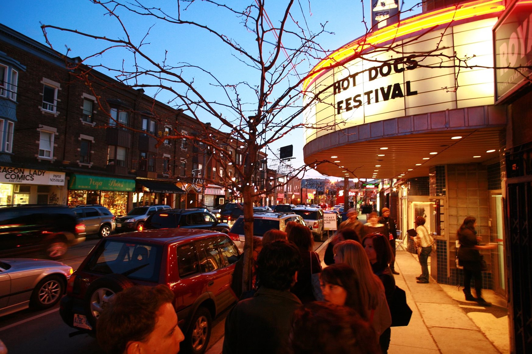 Exterior of a cinema with marquee reading Hot Docs and a line up of people at dusk.