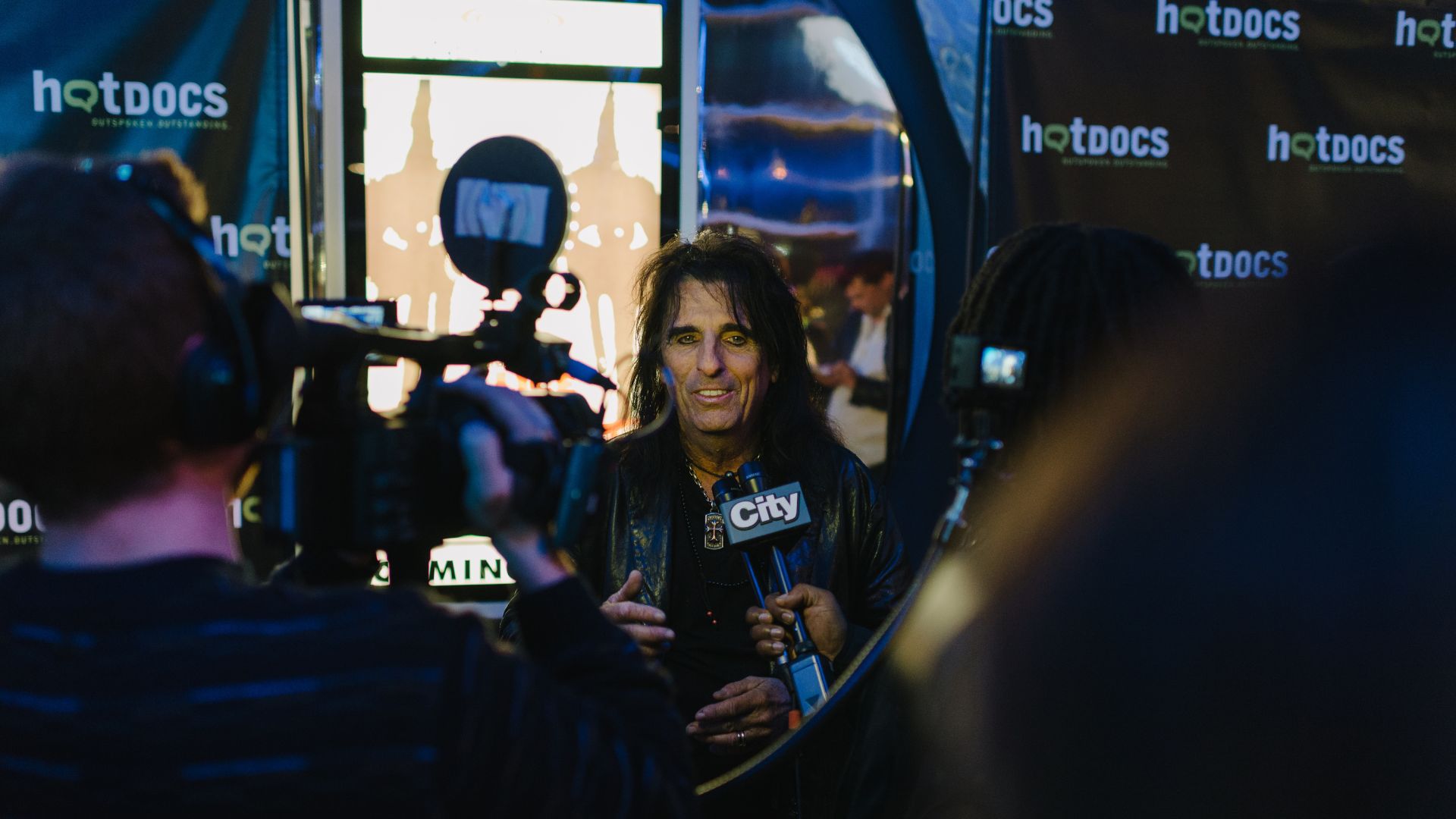 Alice Cooper speaking into a City News microphone surrounded by journalists outdoors with Hot Docs logo displayed in the background.